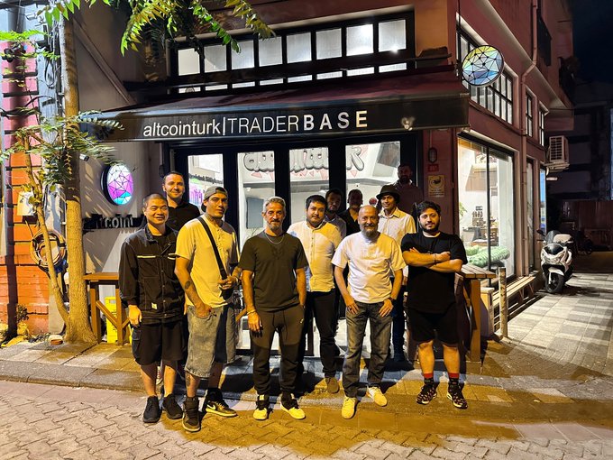 A group of men pose for a photo at night on a city street in front of a storefront with the sign 'altcointurk | TRADER BASE'.