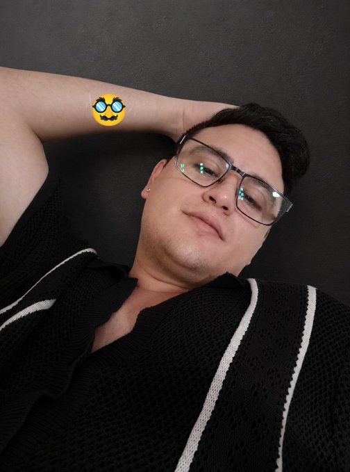 A man wearing glasses and a black knit vest with white stripes poses against a dark wall, one arm raised behind his head in a relaxed stance. A skull emoji with blue glowing eyes is overlaid on his arm, adding a playful, edgy touch that complements his casual GM (good morning) post about taking it easy and feeling ready to bury more NFTs on ETH.
