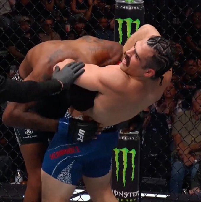 A UFC fighter executes a choke submission on his opponent against the cage, with visible "Monster Energy," "Crypto.com," and "UFC" sponsorship branding, capturing the moment described where Luis Hernandez choked out Sedriques Dumas in his debut.