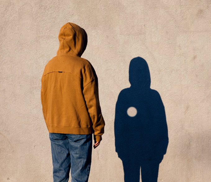 The image features a person wearing a brown hoodie, standing against a wall with their shadow cast onto the wall. The person's shadow appears to be of another individual who has a heart-shaped patch on their chest. The background of the image is white, which contrasts with the brown hoodie and the blue shadow of the other person.