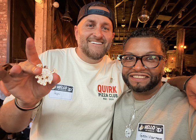 Two smiling men wearing 'HELLO MY NAME IS' stickers and shirts reading 'QUIRK PIZZA CLUB' at what appears to be a networking event.