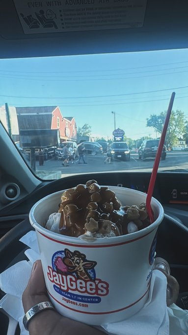 A hand wearing a silver ring holds a cup of frozen custard topped with caramel from 'JayGee's' (jaygees.com, Heart of it Center) inside a car, with a street scene visible through the windshield.