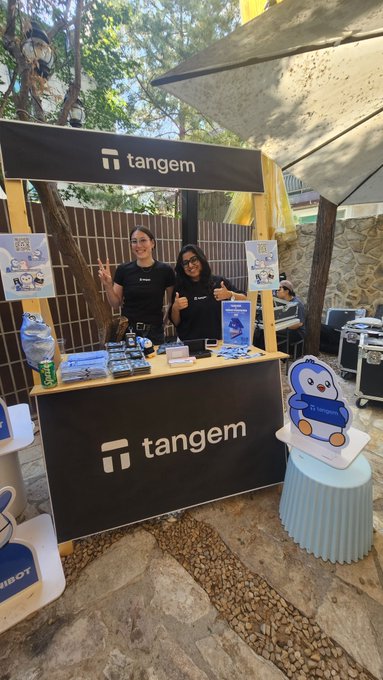 Two people pose at a 'tangem' branded booth with black and yellow signage, where Tangem merchandise and a penguin mascot cutout are displayed.