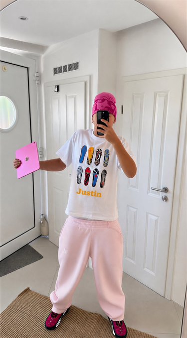 A person takes a mirror selfie wearing a pink head wrap, a white t-shirt with colorful shoe graphics and 'Justin' printed in yellow, pink sweatpants, and pink sneakers, while holding a pink folder.