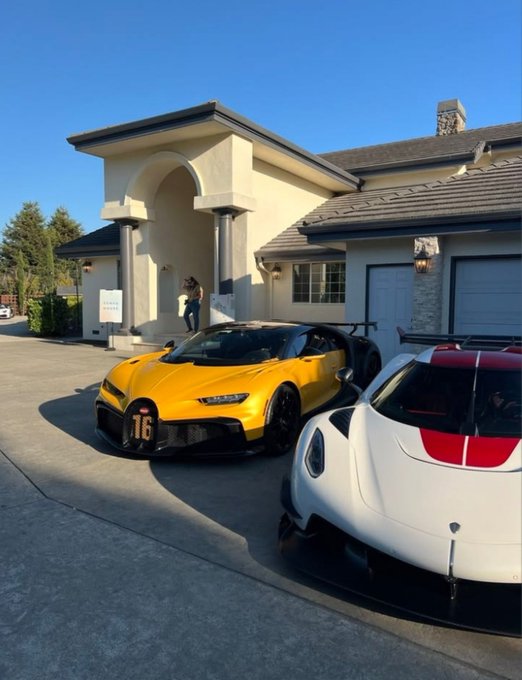 A yellow Bugatti Chiron (numbered 16) and a white sports car are parked in the driveway of a large beige house.