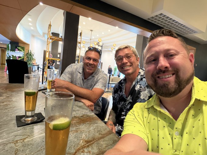 A selfie of three men smiling at a bar, with one taking the photo in a bright yellow shirt, and beer glasses with lime wedges on the marble counter.