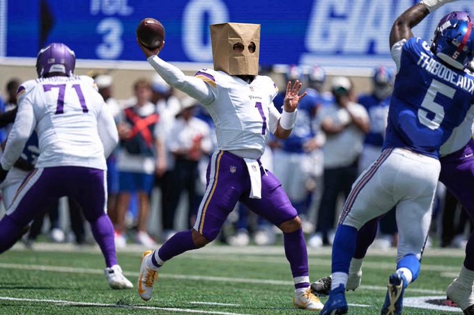 An American football player wearing a Minnesota Vikings white and purple uniform with the number 1 throws a football while wearing a brown paper bag over his head, with other players in white and blue uniforms nearby.