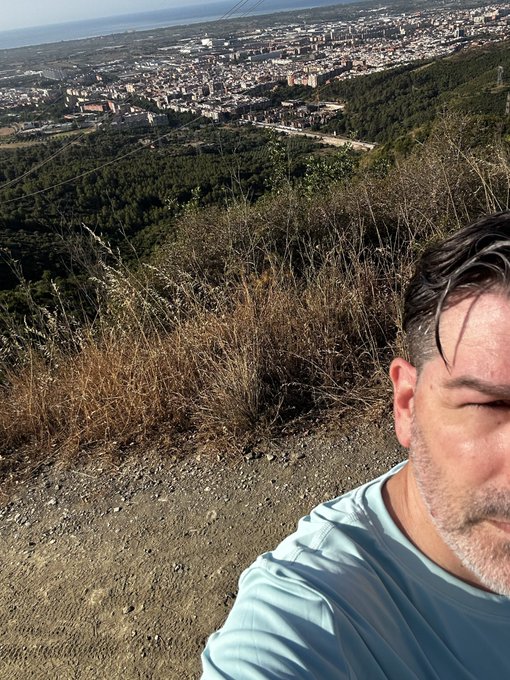 A selfie of a man in a light blue shirt taken on a hillside overlooking a sprawling coastal city near the ocean.