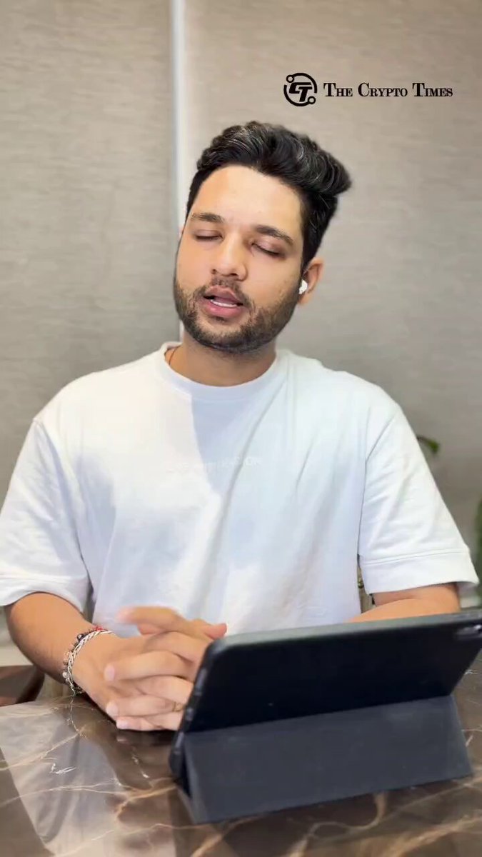 The image shows a man sitting at a table with his hands clasped together while looking down at a tablet computer. The man appears to be wearing a white shirt, which contrasts with the dark background of the photo. There are no other people or objects in the image besides the man and the tablet.