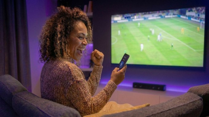 The image shows a woman sitting comfortably on a couch with her arms raised while holding a remote control. She appears to be watching television or playing a video game using a Wii controller. The scene suggests that she might be enjoying some leisure time, possibly engaging in an interactive gaming experience or watching a sports event like soccer.