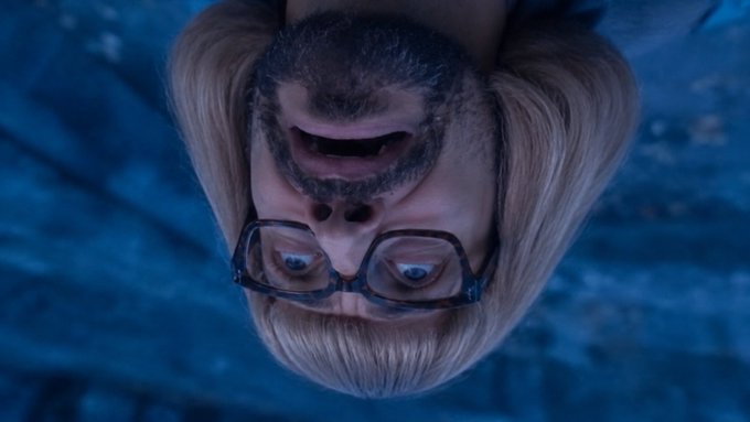 An upside-down cinematic shot of a blonde, bearded man wearing glasses, appearing distressed against a dark blue background.