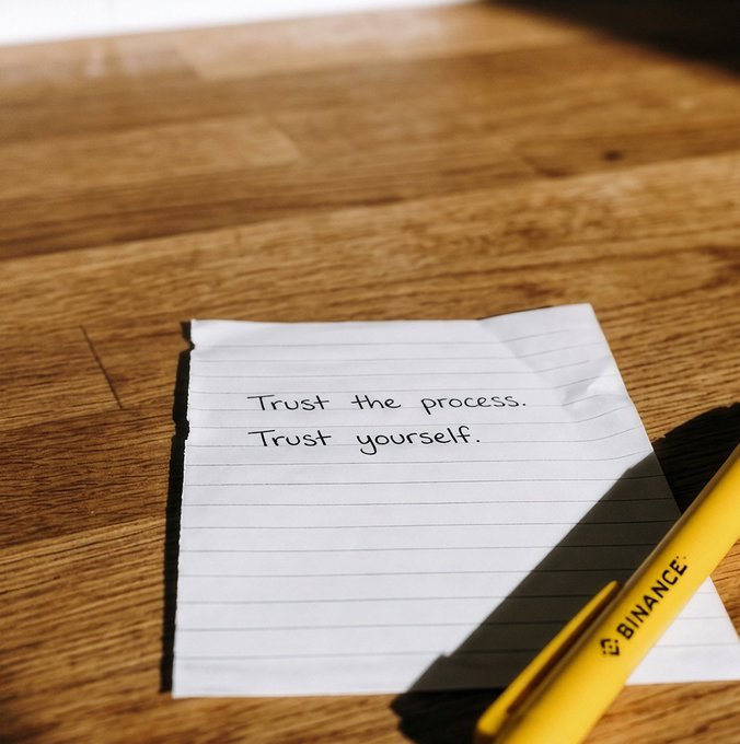 A yellow Binance pen rests on a lined note on a wooden surface, with the handwritten message "Trust the process. Trust yourself." The simple, uplifting note perfectly complements the post's encouraging tone to start the week on a positive note.