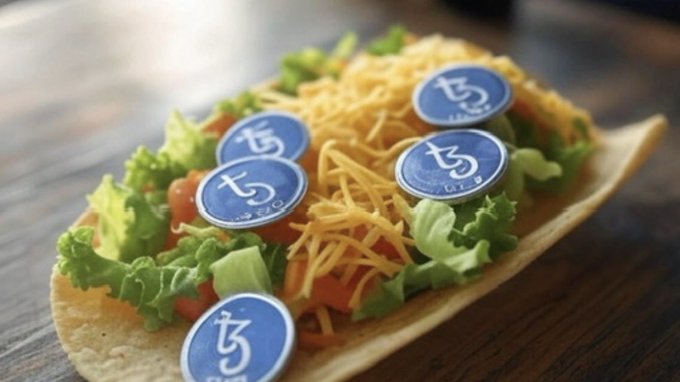 The image features a plate of food with cheese, lettuce, and tomato arranged on top of it. There are six blue buttons placed around the edge of the plate, which appear to be part of an art project or promotional activity related to tezos (Japanese yen). The text "drop your tezos art!!! Is tezos Tuesday!!!", written in white letters, suggests that this could be a social media post promoting tezos or an event involving tezos.