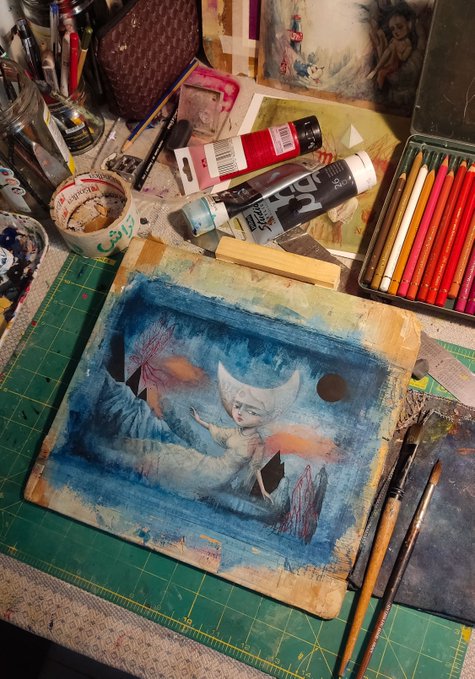 An artist's workspace featuring a watercolor painting of a moon-faced figure reclining, surrounded by paint tubes, brushes, colored pencils, and art supplies on a cutting mat.