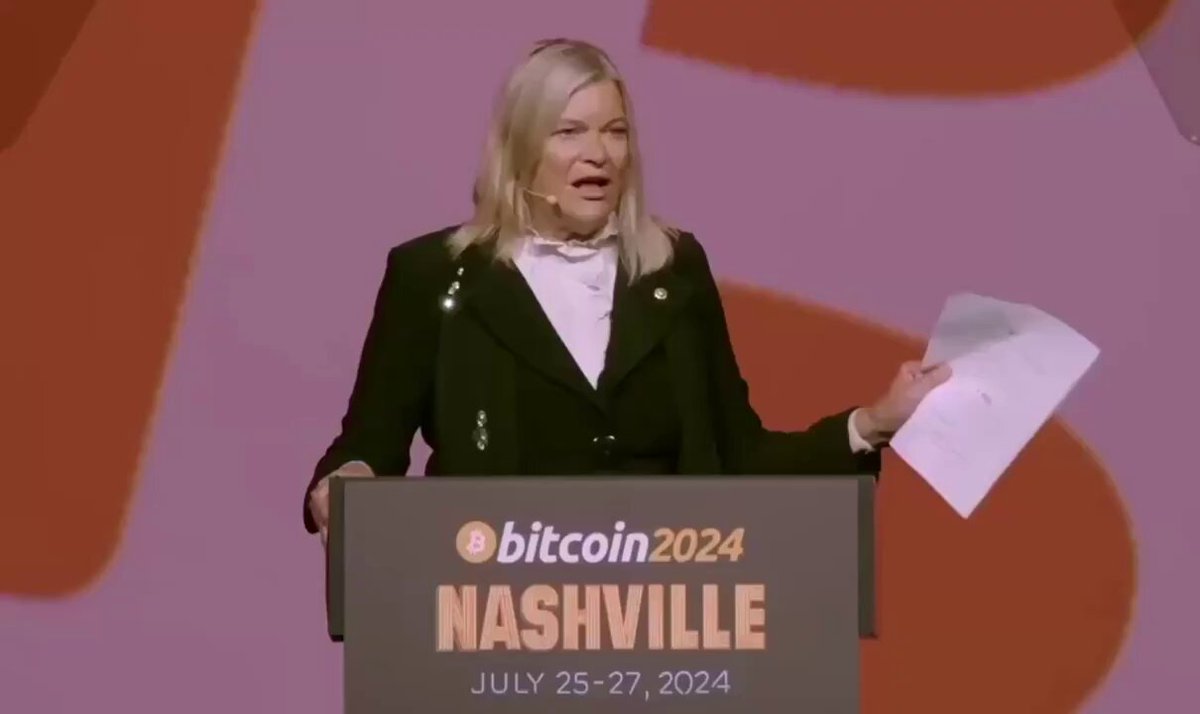 The image shows a woman standing at a podium on stage with a large poster behind her, which reads "BITCOIN2024 NASHVILLE JUL 25 - 27 2014". The background of the stage features an orange and pink patterned backdrop. The woman appears to be addressing a crowd or giving a speech related to Bitcoin and its potential impact on various aspects, including the economy, technology, and financial markets. The post suggests that she is speaking about Bitcoin's future value and its relevance in today's rapidly changing landscape of digital currency.