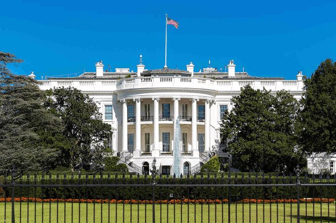 The White House stands prominently under a clear blue sky, with the American flag flying above its iconic columned façade and a fountain visible on the South Lawn. The image connects to the post by symbolizing the executive branch's role in formalizing the U.S. Strategic Bitcoin Reserve, underscoring the administration's involvement in establishing the U.S. as the largest government holder of Bitcoin.