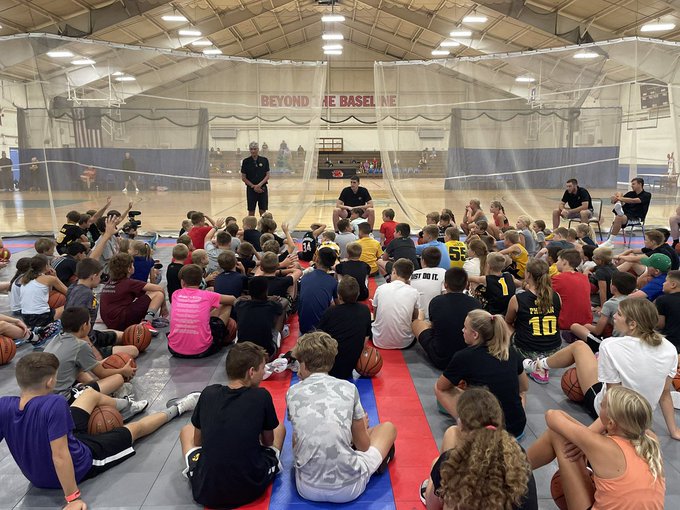 A large group of young basketball campers sits attentively on the gym floor, listening to an instructor with a banner reading "BEYOND THE BASELINE" hanging in the background, as part of a six-day countdown to Luka Garza's motivational basketball camp.