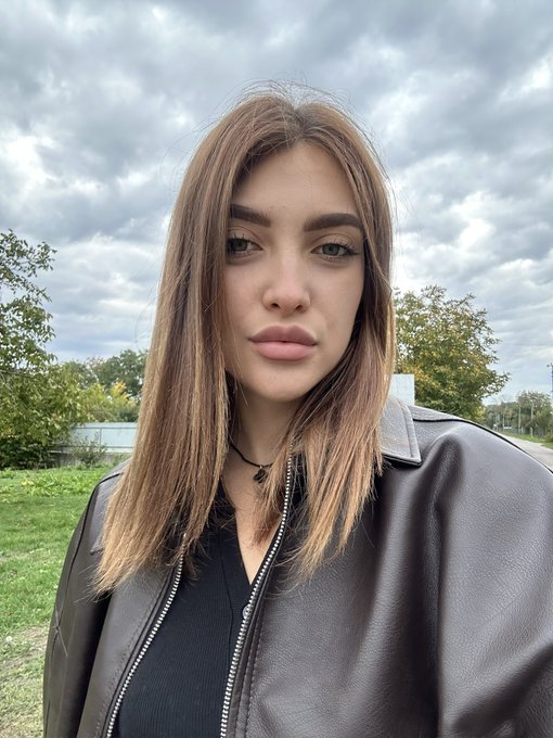Selfie of a young woman with long brown hair wearing a dark leather jacket and necklace, standing outdoors against a cloudy sky.