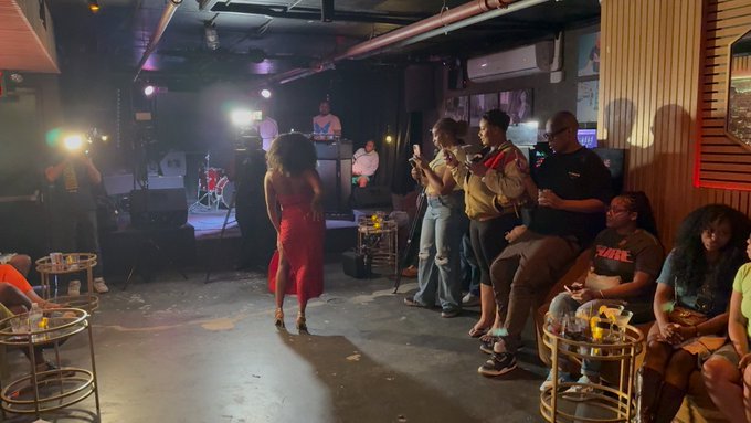 A live music venue scene with a woman in a red dress on stage, audience members standing and sitting, and a band performing in the background.