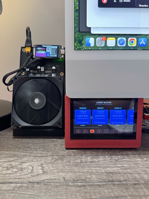A Bitcoin mining setup featuring a NerdMiner device with a cooling fan next to a SoloSatoshi unit displaying 'LATEST BLOCKS' with block numbers 966842, 966841, and 966840, positioned in front of an iPad.