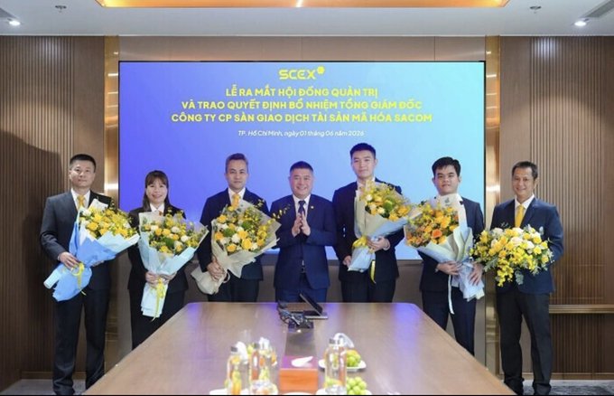 A group of eight people in business attire pose holding bouquets of yellow flowers in front of a screen displaying Vietnamese text including "SCEX," "LỄ RA MẮT HỘI ĐỒNG QUẢN TRỊ," and "CÔNG TY CP SẢN XUẤT DỊCH TÀI SẢN MA HOA SACOM."