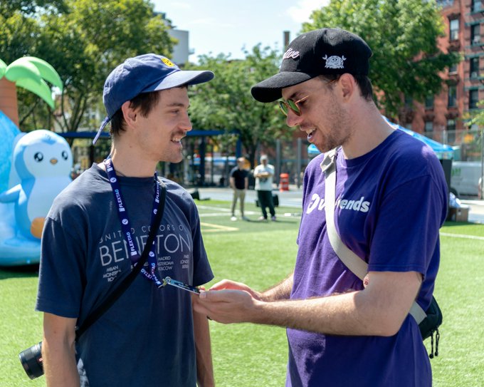 Two young men smiling and looking at something together at an outdoor event, one wearing a blue cap and 'Beffton' shirt, the other in a black cap and purple 'Doginals' shirt.