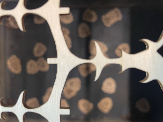 A close-up of a silver metallic cutout design with intricate pointed and curved shapes, with blurred brown stone-like objects visible through and behind the openwork pattern.
