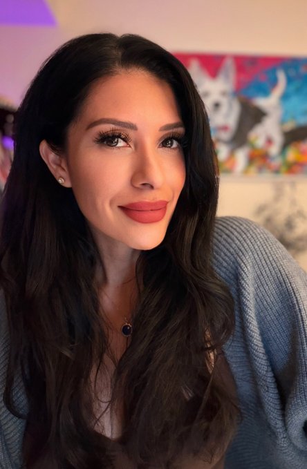 A young woman with long dark wavy hair, wearing a blue cardigan, coral lipstick, and a dark pendant necklace, smiles warmly at the camera in what appears to be a cozy, art-filled room. Her polished, ready-for-the-day look matches the upbeat Monday morning energy of the post, projecting the focused and motivated attitude of someone "locked in and ready to cook" for a productive week ahead.