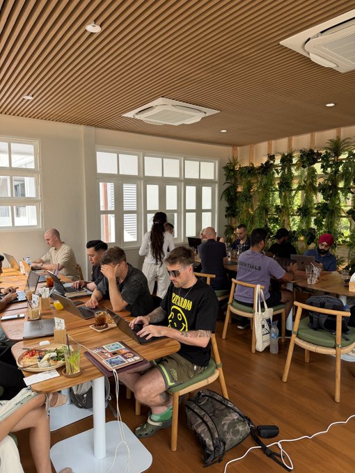 A bustling cafe or co-working space with wood-slatted ceilings where several patrons sit at long wooden tables working on laptops, with a man in a black 'DRAGON' t-shirt and sunglasses typing in the foreground.