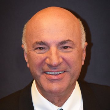 A close-up portrait of Kevin O'Leary, the entrepreneur and "Shark Tank" personality, smiling warmly against a dark background. The image accompanies his recent remarks comparing crypto to the Labubu toy craze, warning that significant money has been lost in the speculative market.
