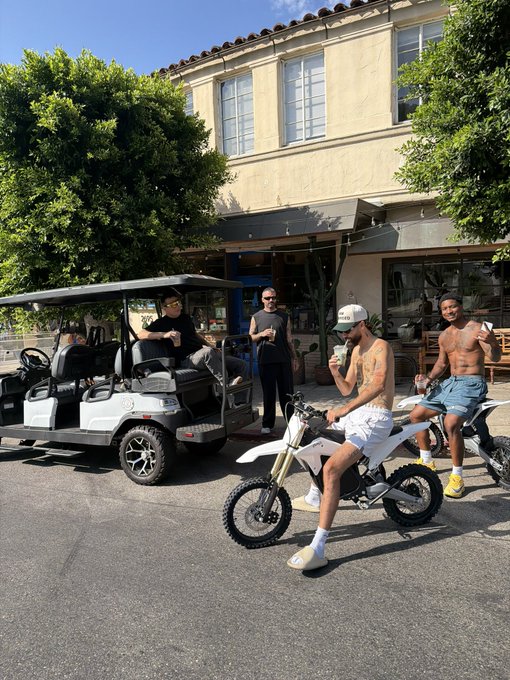 Four young men gather outside a Mediterranean-style building, with one seated on a white dirt bike, another on a matching bike, one behind a golf cart, and a shirtless man in shorts posing casually — capturing a laid-back, sunny "SOLANA SUMMER" vibe. The relaxed street scene, surfboards, and casual attire embody the easy, warm-weather energy suggested by the post.