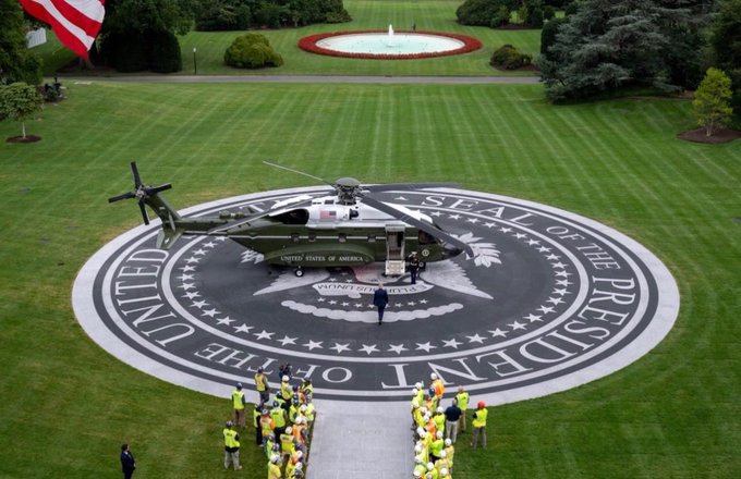 An aerial shot shows the Marine One helicopter landed on the Seal of the President of the United States emblem on the White House lawn, with workers in yellow vests gathered nearby.