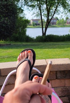 A first-person view of feet in flip-flops resting on a low brick wall by a lake, with a hand holding a cigar in the foreground.
