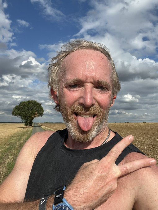 A selfie of a smiling, sweaty man with a grey beard sticking out his tongue and holding up a peace sign while running outdoors on a country road under a partly cloudy sky, wearing a black tank top and a Garmin watch.