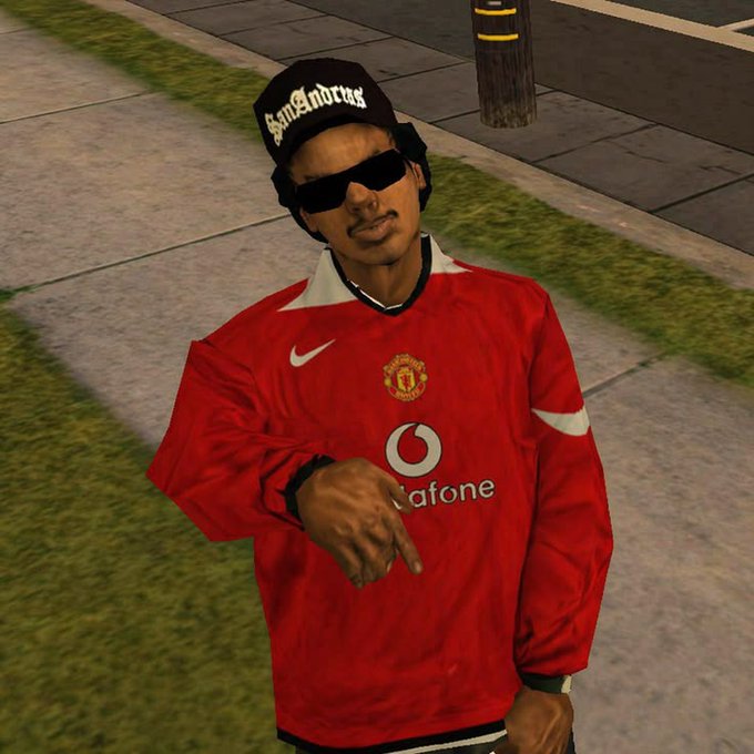 The image features a man wearing a red shirt with "Man City" written across the front, standing on a sidewalk next to a street. He has sunglasses on his head and is pointing at something in the distance. The surrounding area includes grass and a sidewalk, giving an impression of an urban setting. There are also two cars visible in the background, further emphasizing that this scene takes place in a city environment.