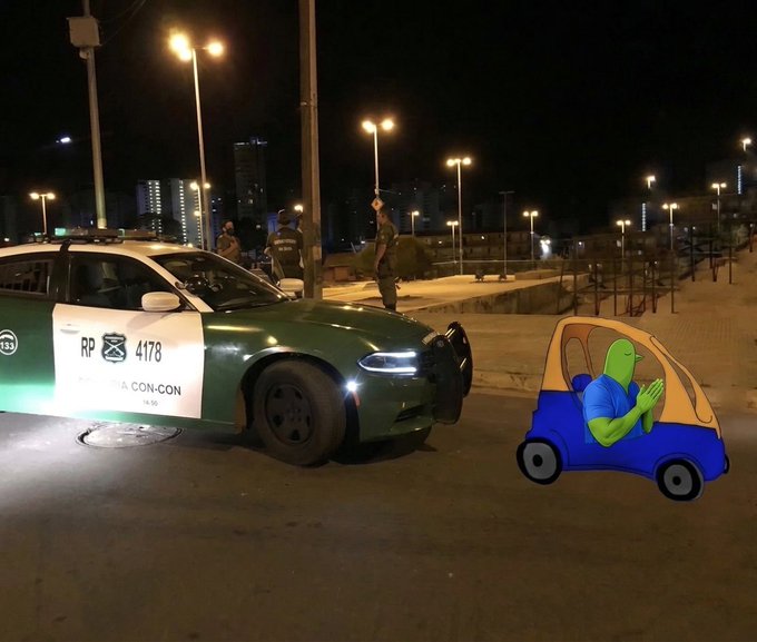 Nighttime street scene with a green police car and officers, featuring a cartoon Pepe-the-Frog character composited into a tiny blue car on the right side.