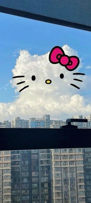 A photograph taken from a window showing a large cloud formation that has been edited with a pink Hello Kitty bow and face, with city buildings visible below.