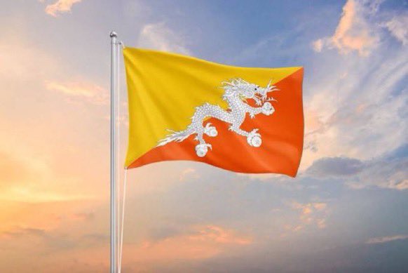 The national flag of Bhutan, featuring a yellow and orange diagonal half with a white dragon, waving against a sunset sky.