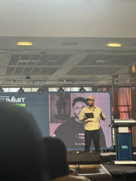 A view from the audience at a conference showing a speaker on stage with 'THE SUMMIT' displayed on a large screen, with the silhouette of an audience member's head visible in the foreground.