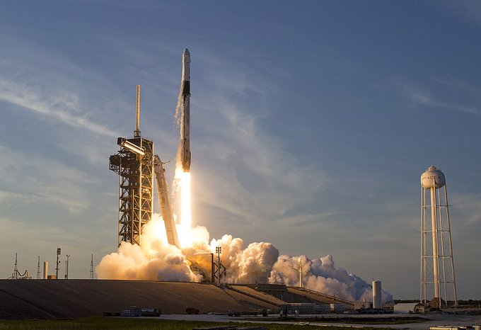 A photograph of a SpaceX Falcon 9 rocket launching from a pad, with bright flames and smoke billowing out, set against a blue sky with thin clouds.