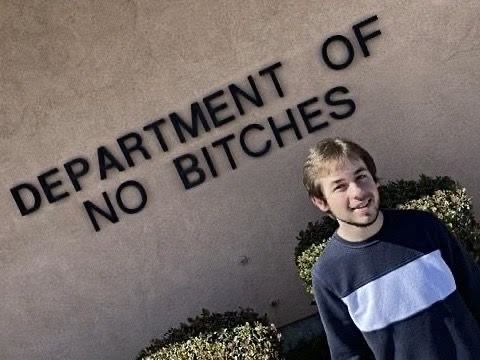 A photo of a man smiling next to a wall sign that reads 'DEPARTMENT OF NO BITCHES'.