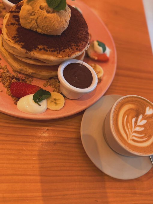 A stack of pancakes topped with cream and mint, served alongside fresh strawberries, banana, and a small pot of chocolate sauce on a coral plate, with a beautifully latte-art cappuccino on the wooden table beside it. The cozy brunch scene ties into the post's caption about seamlessly paying with a Nexo crypto card, illustrating how everyday indulgences can be enjoyed effortlessly through crypto transactions.