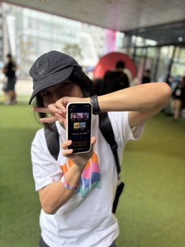 A young person wearing a black bucket hat and white t-shirt with a colorful logo takes a photo using a smartphone at what appears to be an outdoor event with green turf flooring.
