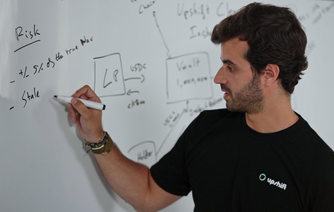 A man wearing an Upshift-branded black t-shirt writes risk-related notes (including "+/- 5% of the true NAV," "Stale," and LP/Vault diagrams) on a whiteboard.