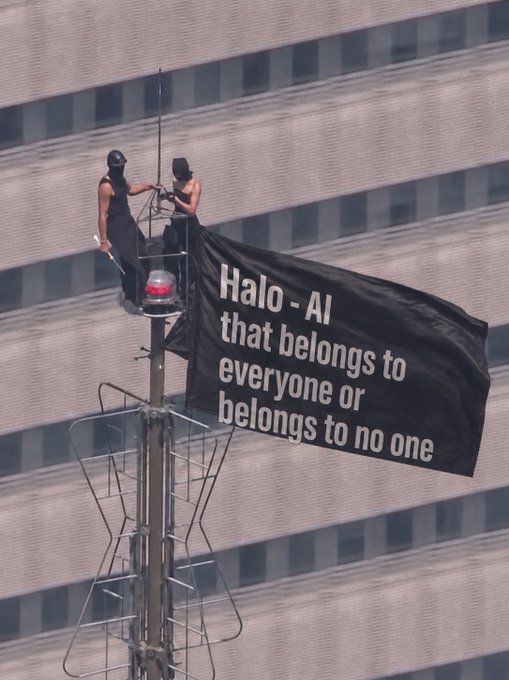 Two activists stand atop a communications tower, unfurling a large black banner that reads "Halo - AI that belongs to everyone or belongs to no one," directly delivering the protest's message against corporate AI monopolization in a highly visible urban setting. The dramatic aerial placement of the banner emphasizes the activists' call for AI to remain a shared public resource rather than be controlled by any single entity.