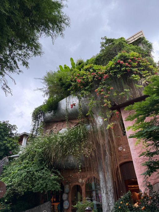 A pink, ornate building overgrown with lush greenery, vines, and pink flowers under a cloudy sky.