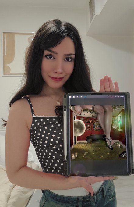 A young woman with long dark hair, wearing a black and white polka-dot top, holds up a tablet displaying a digital artwork of legs in heels sitting above a pan of cooking rice.