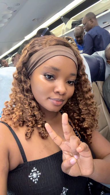 A woman with curly brown hair wearing a brown headband and a black patterned top takes a selfie with a peace sign while seated on an airplane.