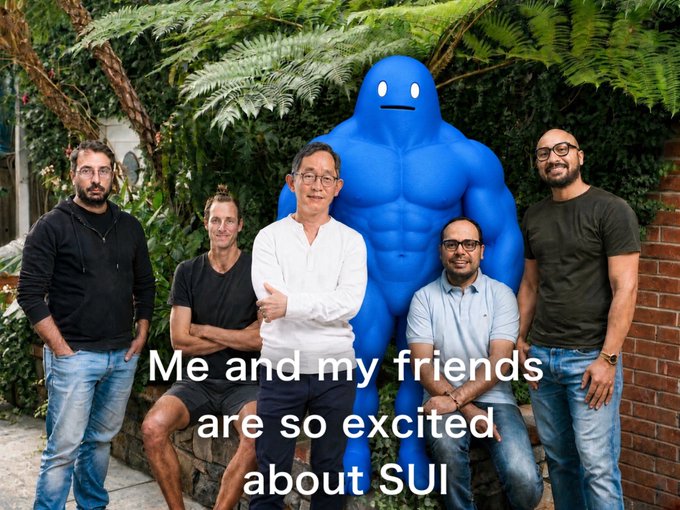 Group photo of five men standing in front of greenery with a large blue cartoon mascot character behind them, overlaid with white text reading "Me and my friends are so excited about SUI" with the SUI logo.