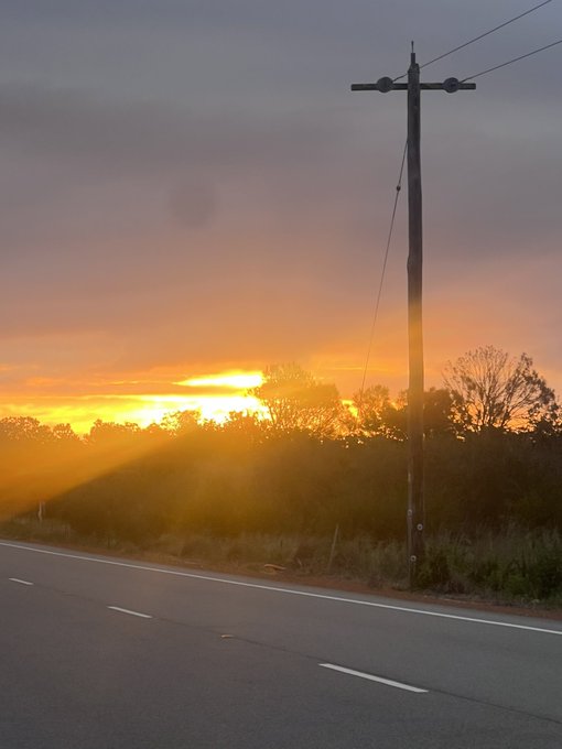 A scenic photograph of a sunset with vibrant orange and yellow rays streaming over a landscape, with a utility pole silhouetted in the foreground beside a road.
